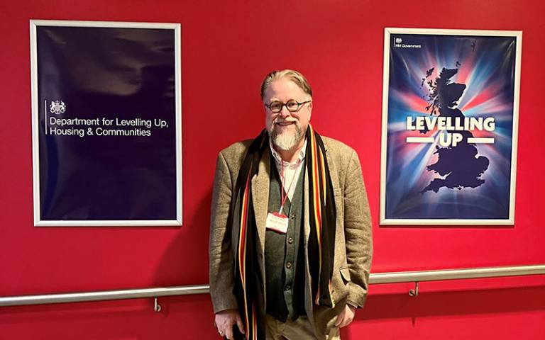 Prof Kevin MacDonald (Director, UCL Institute of Archaeology) presented the Black History Month Lecture to UK Government Departments (Oct 2022). Kevin is standing in front of a red background, between two posters of the Department for Levelling Up