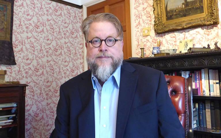 Bearded man with glasses wearing a navy suit jackey and light shirt sitting in a room with patterned wallpaper and paintings and a bookcase behind him