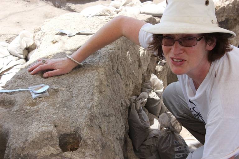 Woman wearung a sunhat, sunglasses and lightcoloured t-shirt and trousers working on an archaeological site, kneeling and leaning against the side of a trench