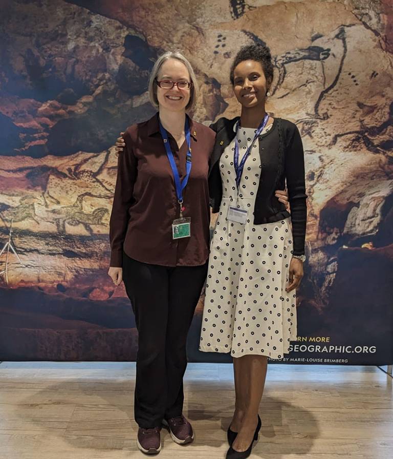Two women standing together in front of a picture/banner of rock art paintings 