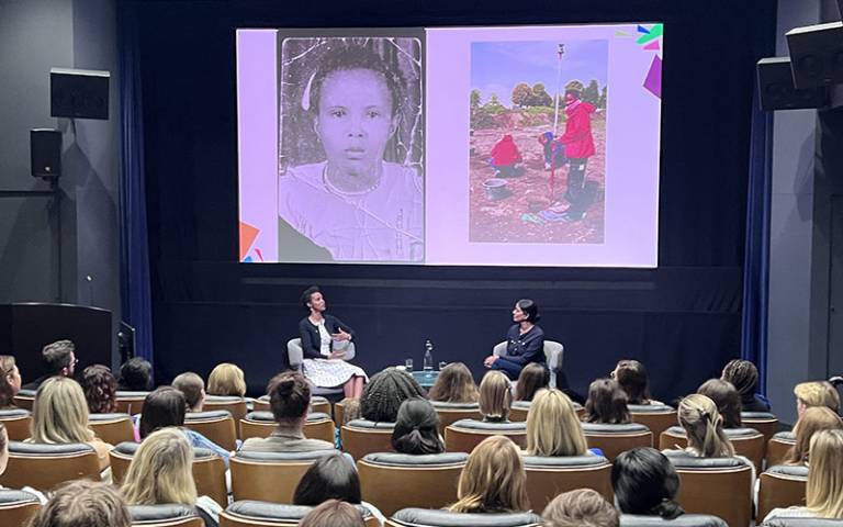 Two women sitting facing each other on a stage with a screen in the background showing the image of a young girl and also a young woman undertaking fieldwork