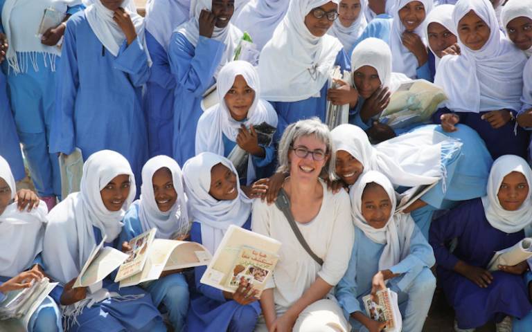 Woman (Dr Claudia Naeser, wearing glasses and white clothing) in centre surrounded by a group of girls, dressed in blue clothing with white headscarves (uniform), holding books
