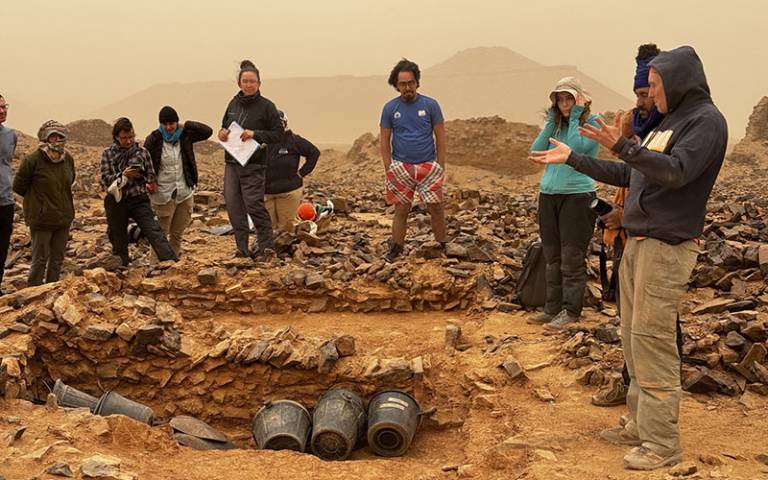A man (Giles, UCL ASE) presenting a structure to other people standing around the edge of an archaeological trench in a desert location