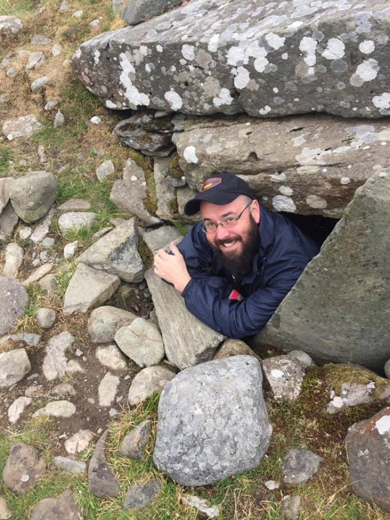 Bearded man, smiling, wearing glasses, a navy raincoat and black cap looking out from a stone monument (tomb?)