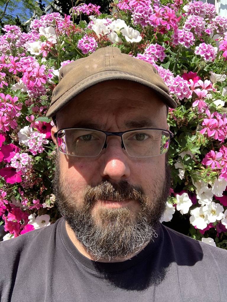 Bearded man wearing glasses, a black top and brown cap, looking into the camera, standing in front of a bush of pink and white flowers