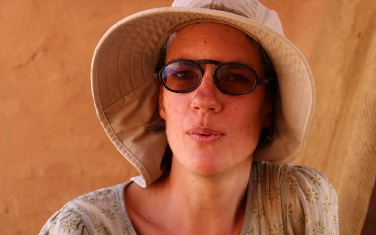 Head and shoulders image of a woman looking directly at the camera wearing sunglasses and a brown sunhat and a blue/grey top with green patterning
