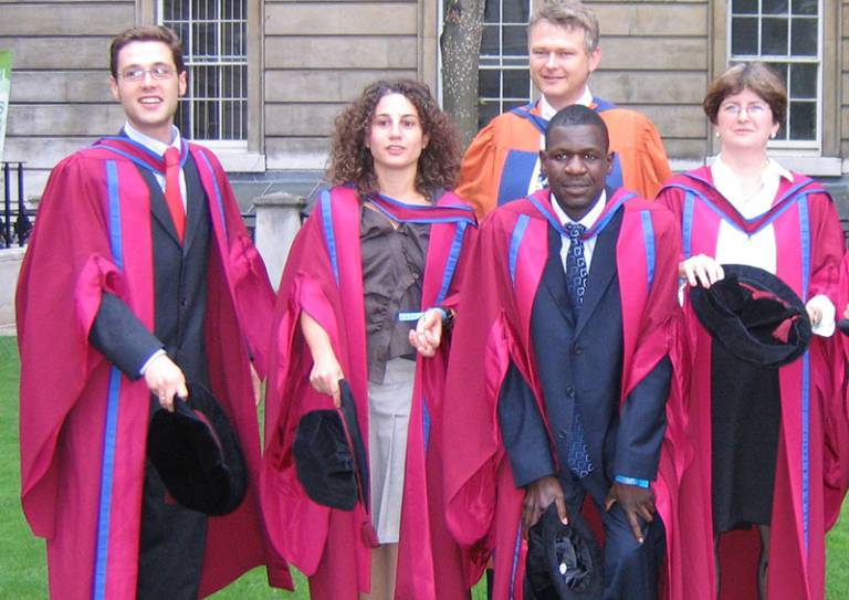 Five people standing outside a building in graduation gowns and hats 