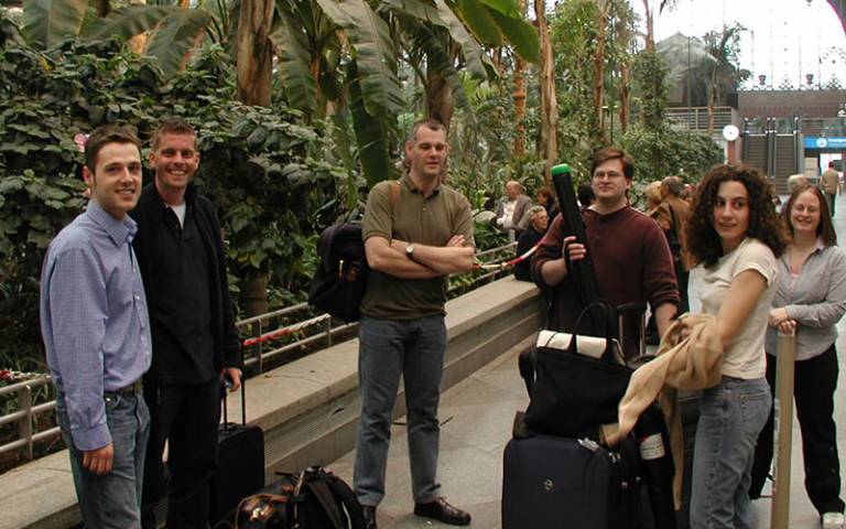 Six people standing together with luggage in an outside area beside palm trees/foliage