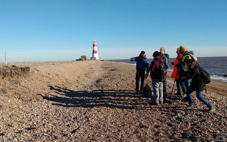 Group of people filming in a shingle beach location with a white and red lighthouse in the background