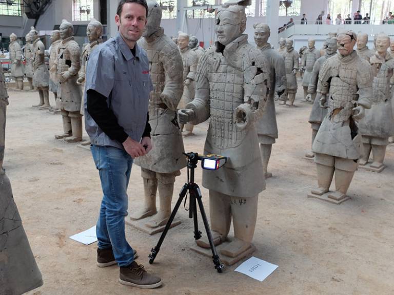 Man, wearing a grey top and jeans, looking at the camera, standing next to a Terracotta Army figure 