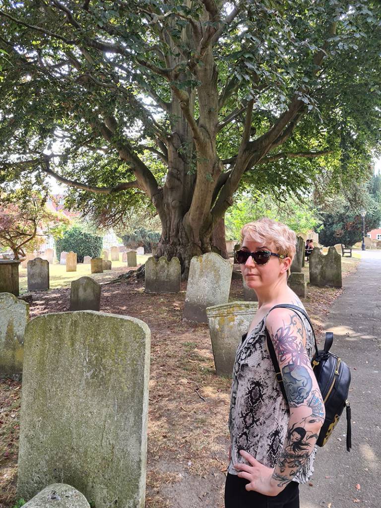 Woman with blonde hair wearing sunglasses and a B&W top and black rucksack standing on a path in a graveyard setting 