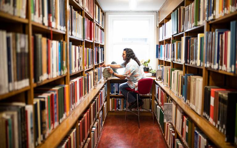 Male student sitting on a high stool at the end of a library corridor surrounded by bookshelves