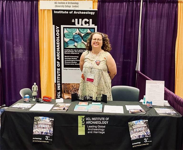 A woman with curly hair and glasses behind a table/stall advertising a university department (purple and orange curtains in the background)