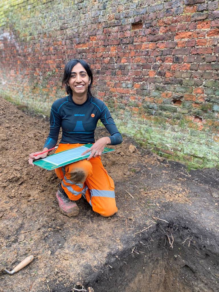 Student in dark blue top and orange high-vis trousers, kneeling on the ground beside the edge of a trench, holding a board on her knee, with a red-bricked wall in the background