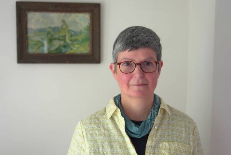 Woman with grey-sh hair and red glasses wearing a yellow and white patterned shirt and green scarf looking at the camera, in front of a light coloured wall with a framed picture in the background