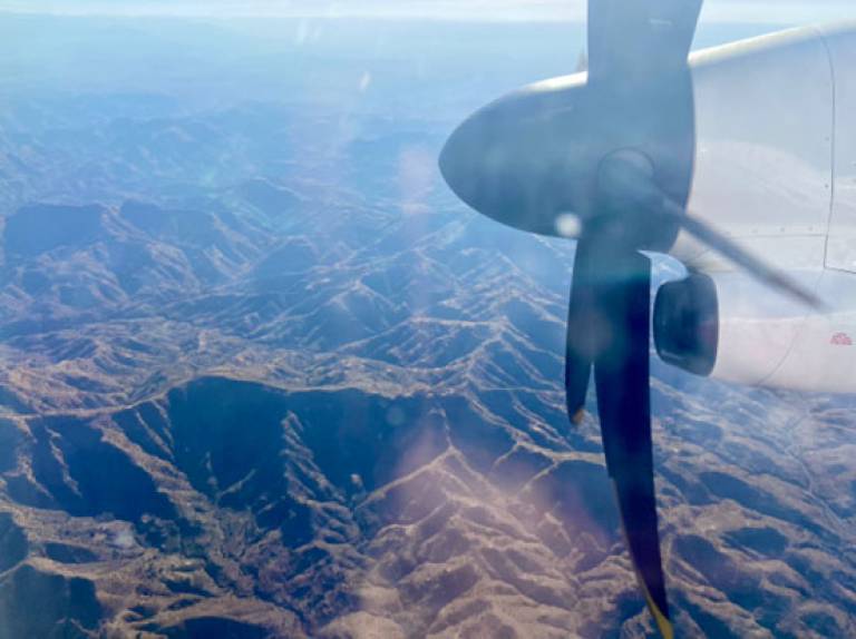 Image from an airplane window of a mountainous landscape below and the plane's engine in the right foreground of the picture
