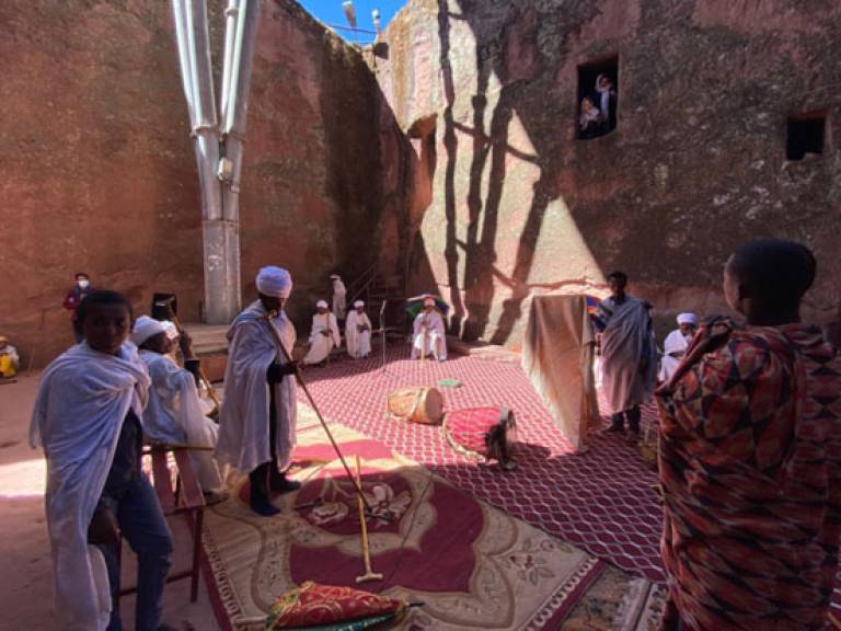 People, some wearing white robes, standing/sitting within a stone structure (with colourful carpets on the floor) conducting a ceremony with onlookers viewing the scene from windows in the walls