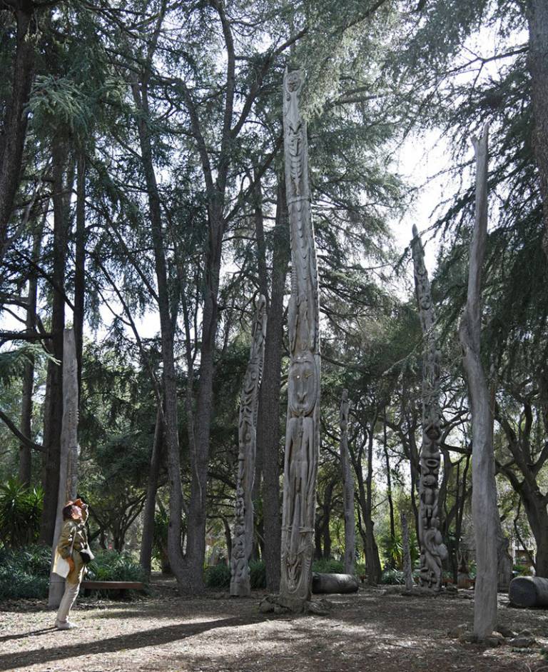 A woman in a wooded area/garden looking up at tall wooden sculptures among the trees 