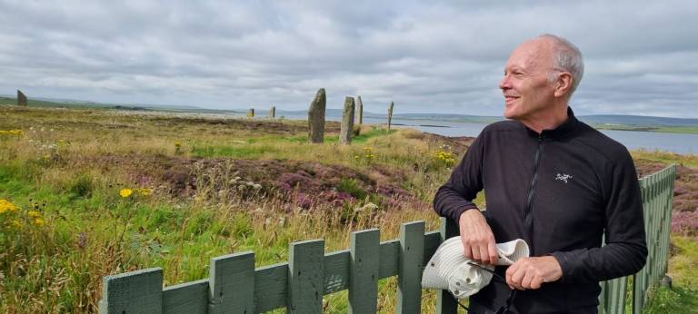 A white-haired man in a black top, standing beside a green fence looking at a stone circle in a field of grass and purple heather