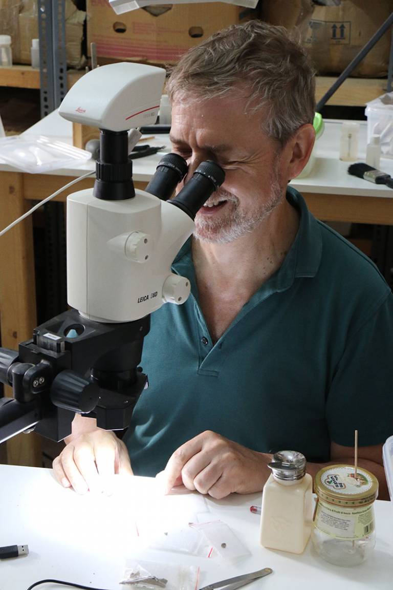 Bearded man, in a green top, smiling, looking down a white microscope