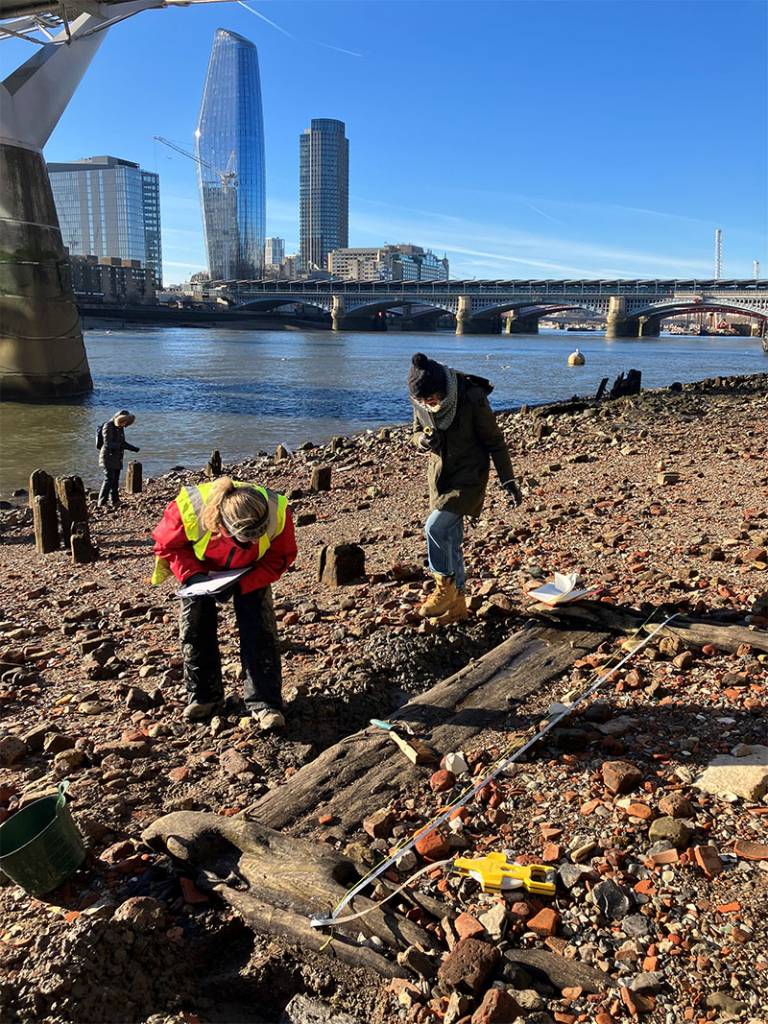 Students wrapped up against teh cold recording wooden remains beside teh River Thames in London on a cold and sunny day with modern buildings and bridges over the river in the background 