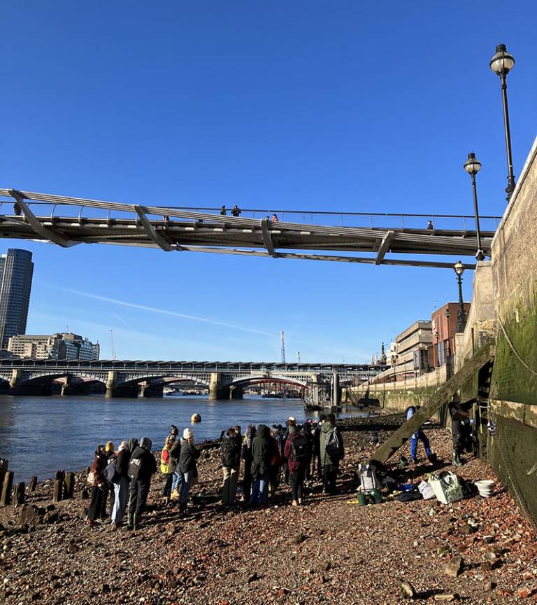 A group of people (students and staff) standing on the side of the River Thames on a cold and sunny day in London with bridges and buildings in the background 