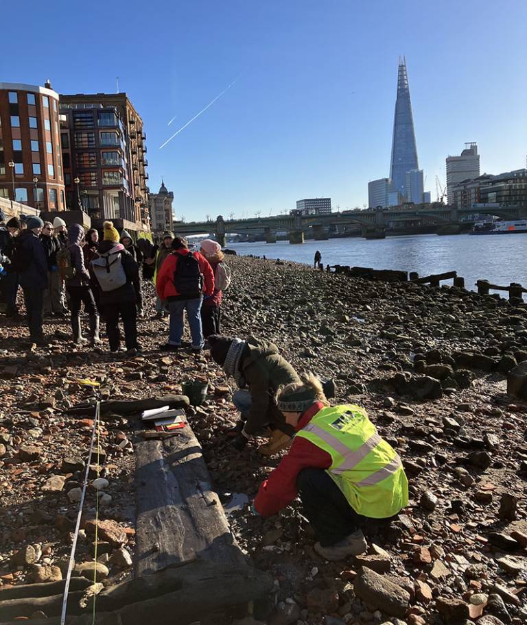 Archaeology students (all wrapped up against the cold) recording wooden remains on the side of the River Thames in London on a sunny day with the Shard building (and blue sky) in the background
