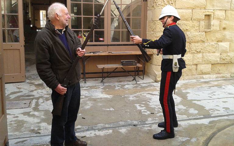 A man in dark clothing laughing with a man in uniform, both holding rifles outside a museum setting
