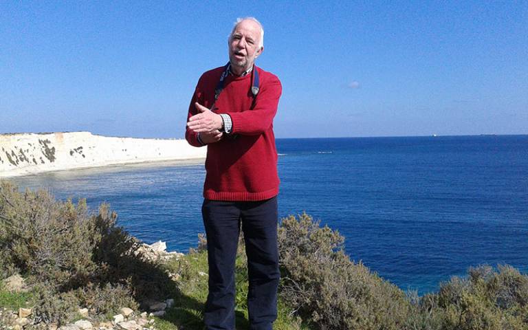 A white haired man wearing a red jumper and dark trousers, holding a camera and standing on a cliff overlooking blue sea and sky