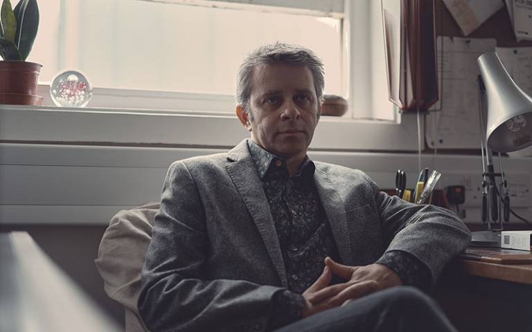 Man, wearing a grey jacket, sitting in a chair in an office setting, in front of a window