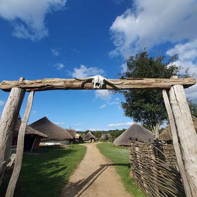 Butser Ancient Farm, Hampshire, UK - a wooden entrance lintel with roundhouses in the background on a sunny day with blue skies overhead