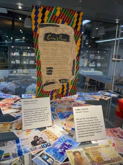A colourful display in an exhibition case including a colourful piece of textile, text panels and currency lying at the bottom of the case