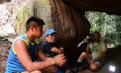 Three men in a cave location with rock art in teh background, one man holding a video camera and interviewing the other men