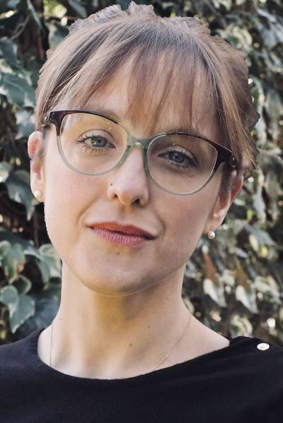 Close up headshot f a woman with brown hair and glasses wearing a black top, standing in front of greenery/foliage