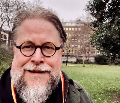 Bearded man with glasses wearing a College scarf in a park/square with a large building in the background