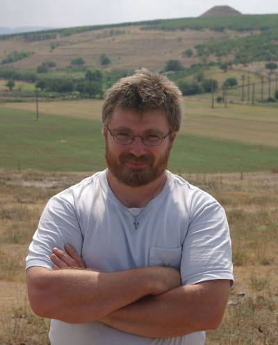 Bearded man with glasses in a white/grey top (Dr Kris Lockyear) standing outside with arms crossed, looking at the camera, with a hill in the background