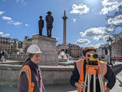 Two archaeologists in high vis jackets stand in front of the iconic statues and fountains of Trafalgar square in bright sunshine. One is looking through a yellow camera-like box (a dumpy level) on a tripod.