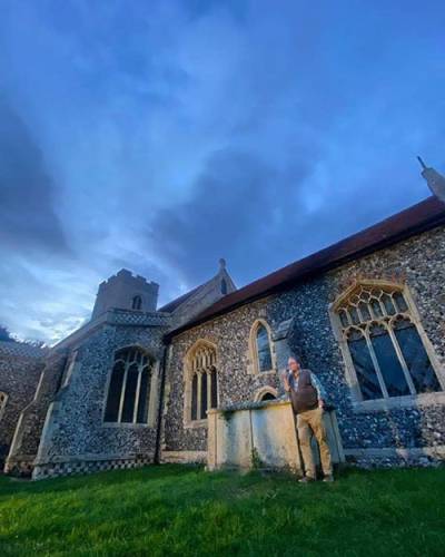 A bearded man smoking a pipe, standing in a churchyard (leaning against a large rectangular tomb) in the dusk
