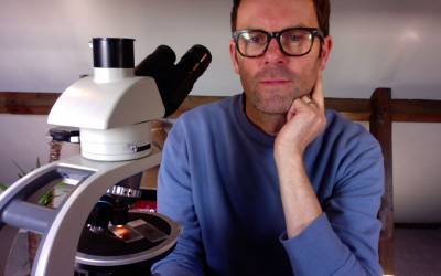 Man in a blue top, wearing glasses, looking at the camera beside a white microscope