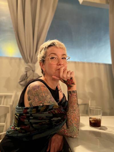 A woman with blonde hair and glasses and tattoos on her arm, hand and shoulder, sitting at a table, wearing a black top and colourful wrap scarf