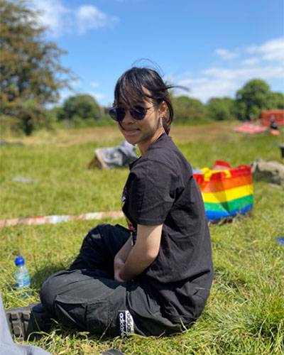 Female student dressed in black, facing the camera side on, sitting on the grass in a field/park location on a sunny day with a rainbow bag behind her 