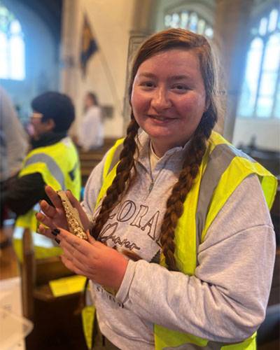 Female student with long brown/red hair in plaits wearing a grey top and yellow high-vis jacket, standing inside a church-like building holding an archaeological find (animal jaw bone)
