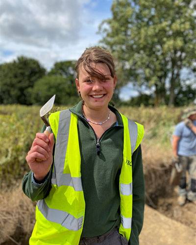 Female student in a green top and yellow high-vis jacket, smiling, standing in a rural location/site and holding up a trowel 