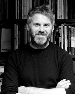 B&W image (head and shoulders) of a bearded man standing in front of a bookcase