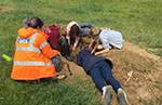 An ASE staff (in orange high-vis jacket) with three young people on an archaeological dig