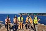 Five people in yellow/orange high-vis clothing, hard hats and harnesses on the top of a cliff overlooking a bay