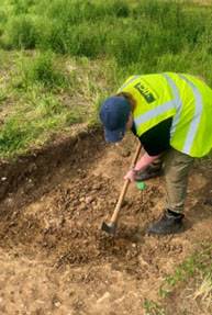A student, wearing a yellow high-vis vest and dark baseball cap, excavating a trench with a mattock during the UCL Norton Fieldschool