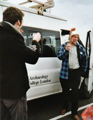 A man in casual clothes wearing a replica Roman helmet standing beside a white minibus having his photo taken