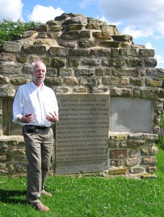 White-haired man in a white shirt and beige trousers standing in front of the remains of a Roman wall which has a plaque inserted in it, on a sunny day