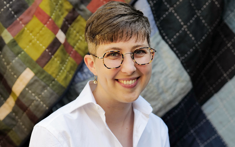 a young woman with short brown hair wearing glasses and a white shirt smiles at the camera. There is a quilt hanging in the background behind her.